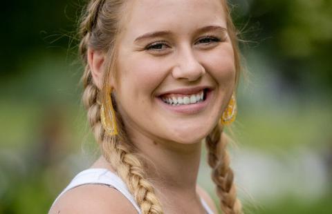 Headshot of Briella Hirsch smiling, wearing Dutch braided pigtails, white tank top, and lemon wedge ear rings.